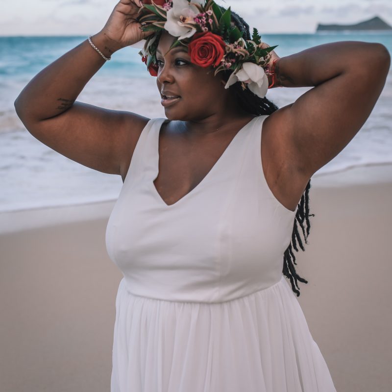 A young Black female stands on a beach, with a crown of tropical flowers on her head